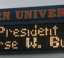An Auburn University sign displays “President George W. Bush” in glowing amber dot-matrix letters against a dark board.