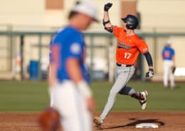 An Auburn baseball player in orange and gray raises a fist while rounding first base, with a blurred Florida defender in front.