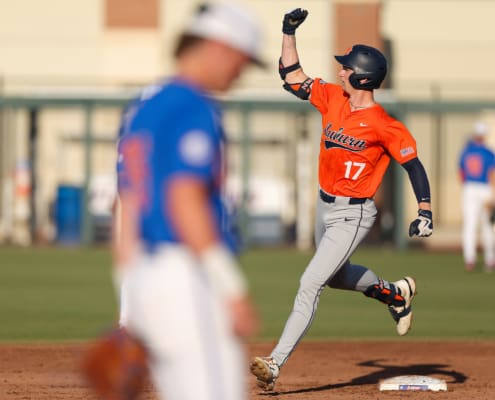An Auburn baseball player in orange and gray raises a fist while rounding first base, with a blurred Florida defender in front.