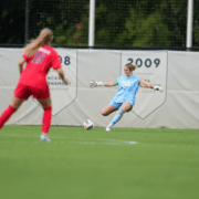 Maddie Prohaska in a light blue Auburn kit swings at a ball while an Ole Miss player in red closes in on the field.