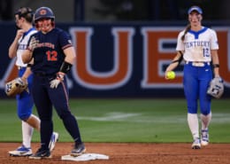 Auburn softball player in navy uniform smiles at second base while Kentucky infielders stand nearby on the diamond.