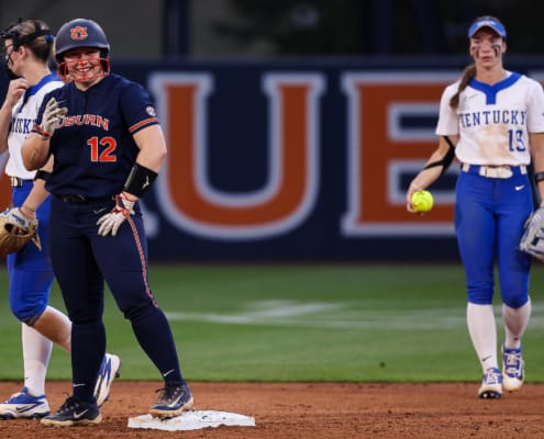 Auburn softball player in navy uniform smiles at second base while Kentucky infielders stand nearby on the diamond.