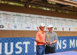 Two golfers pose with an SEC trophy in front of a results board, with college team scores displayed along the back wall.