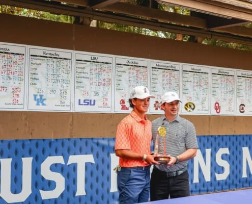Two golfers pose with an SEC trophy in front of a results board, with college team scores displayed along the back wall.