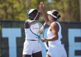 Two Auburn women’s tennis players in white uniforms raise their hands for a high-five on an outdoor court.