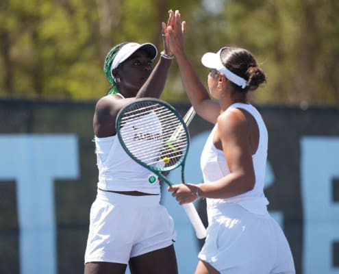 Two Auburn women’s tennis players in white uniforms raise their hands for a high-five on an outdoor court.