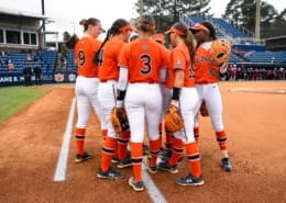 Auburn softball players in orange jerseys huddle on the infield at Jane B. Moore Field under overcast skies.