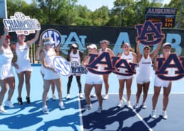 Auburn women’s tennis players celebrate on a bright blue court with SEC championship signs and large Auburn logos.
