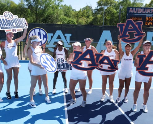 Auburn women’s tennis players celebrate on a bright blue court with SEC championship signs and large Auburn logos.