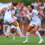 Two Auburn soccer players in white uniforms celebrate on the field, with one grinning as the other jumps nearby.