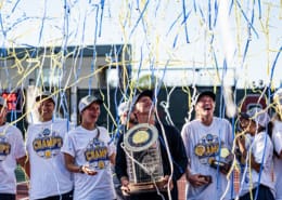 Auburn women's tennis players in white championship shirts smile beneath blue and yellow confetti while holding the SEC trophy.