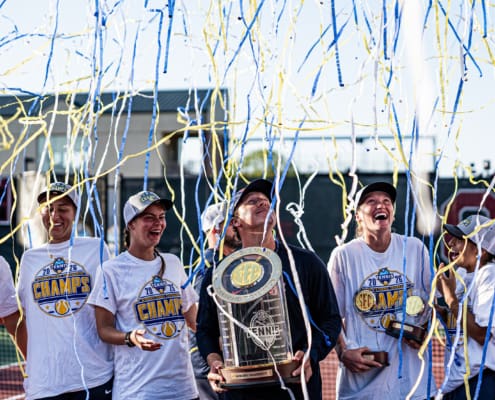 Auburn women's tennis players in white championship shirts smile beneath blue and yellow confetti while holding the SEC trophy.