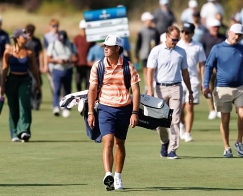 Jackson Koivun walks across a golf course in an orange-striped Auburn polo with a white cap and a bag on his back.