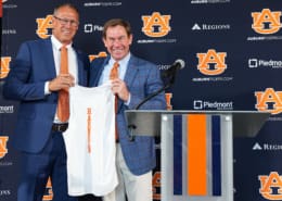 Two men in blue suits hold up a white Auburn shirt beside a podium, with Auburn Tigers logos and sponsor banners behind them.