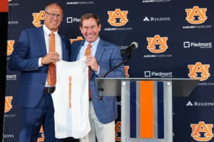 Two men in blue suits hold up a white Auburn shirt beside a podium, with Auburn Tigers logos and sponsor banners behind them.