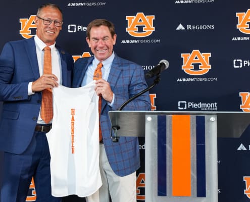 Two men in blue suits hold up a white Auburn shirt beside a podium, with Auburn Tigers logos and sponsor banners behind them.