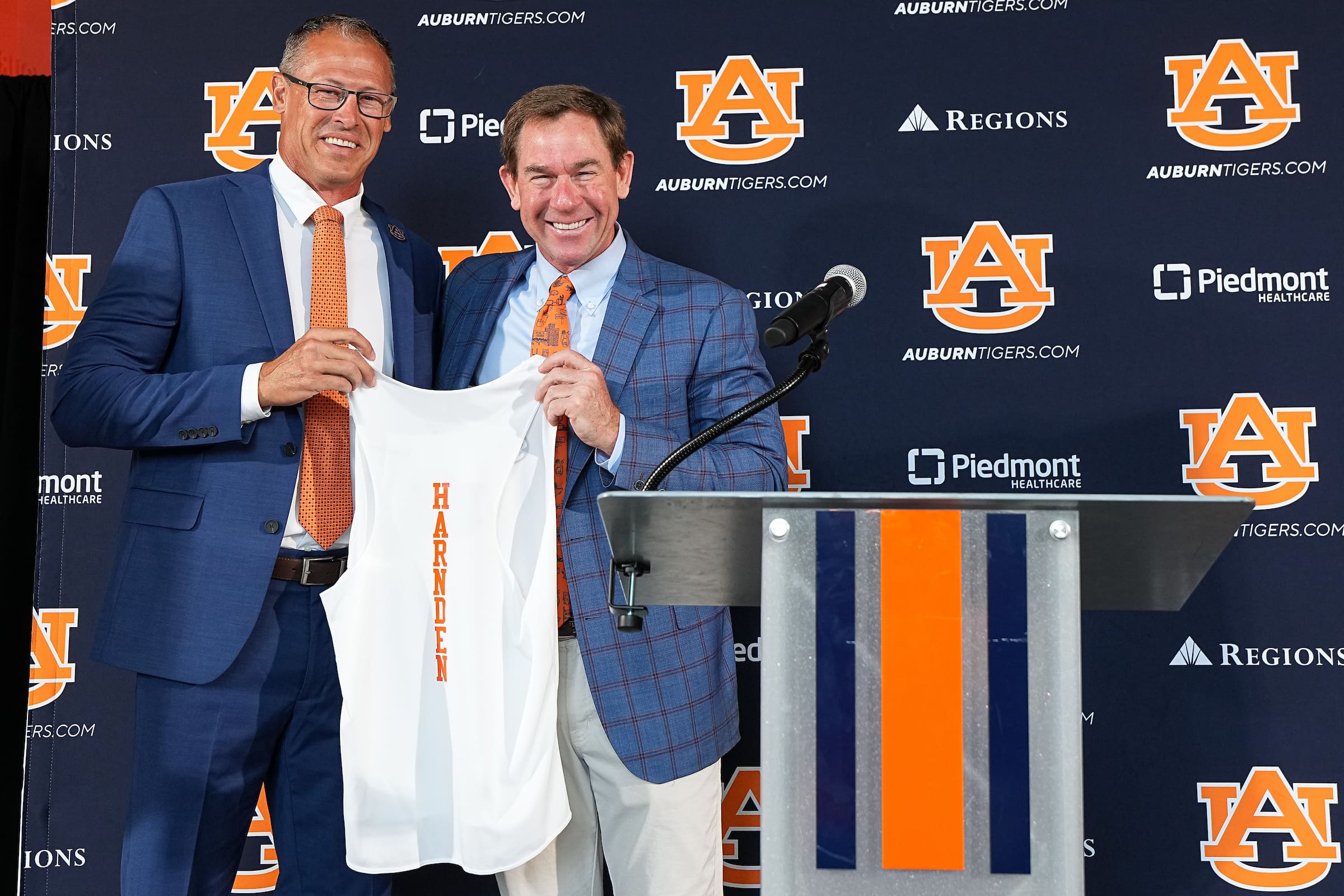 Two men in blue suits hold up a white Auburn shirt beside a podium, with Auburn Tigers logos and sponsor banners behind them.