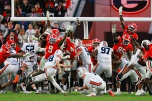 A football hangs above Auburn players in white and Georgia players in red, reaching to block a kick beneath the goalpost.